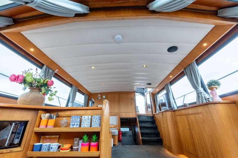 De foto toont het interieur van een boot met houten afwerking, ramen met gordijnen, een trap en diverse decoratieve items.