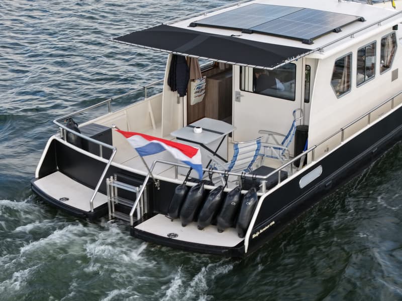 Een witte boot met zwarte romp vaart op het water, met zonnepanelen op het dak en een Nederlandse vlag aan de achtersteven.