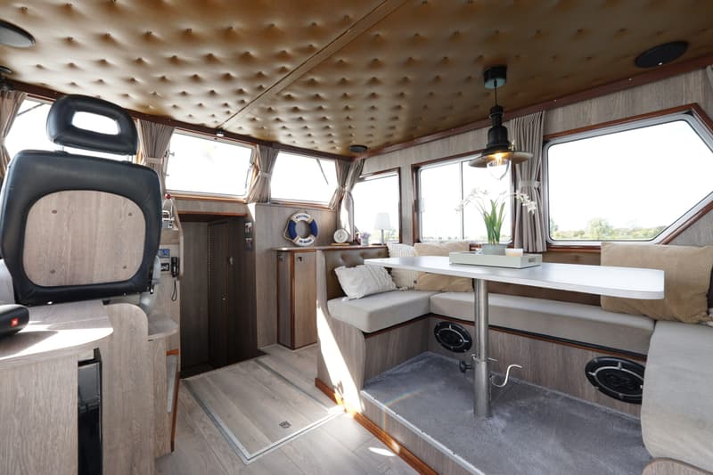 Een interieur van een boot met een stuurstoel, een eettafel met banken, ramen en een gestoffeerd plafond.