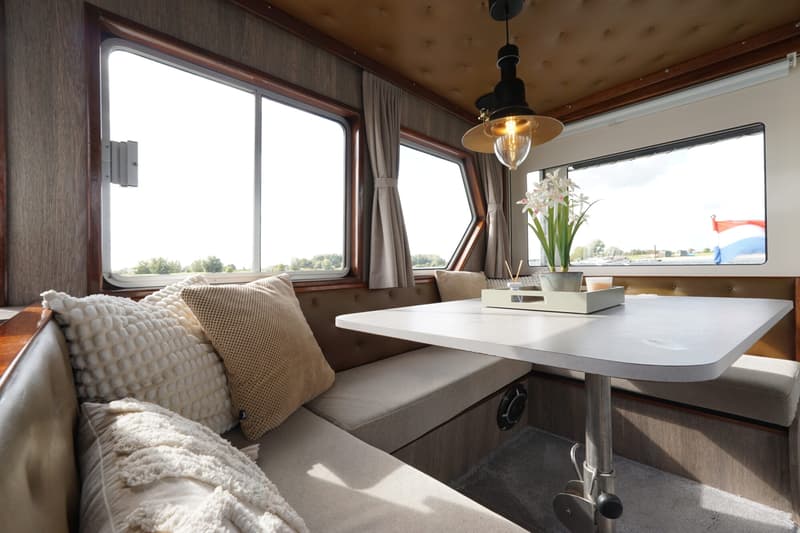 Een interieur van een boot met een tafel, banken met kussens, ramen met uitzicht en een hanglamp.