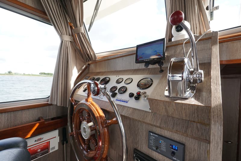 De foto toont het stuurhuis van een boot met een houten stuurwiel, diverse meters, een navigatiescherm en een gashendel.