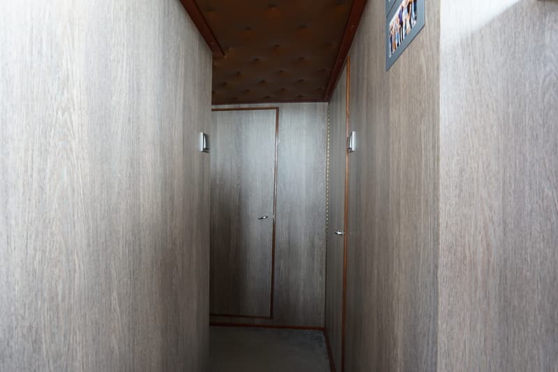Een gang met houten wanden en een bruin, gewatteerd plafond, met twee deuren aan het einde en een aan de rechterkant.