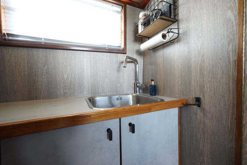De foto toont een kombuis met een rvs-spoelbak, kraan, aanrechtblad, onderkasten en een raam met jaloezieën.