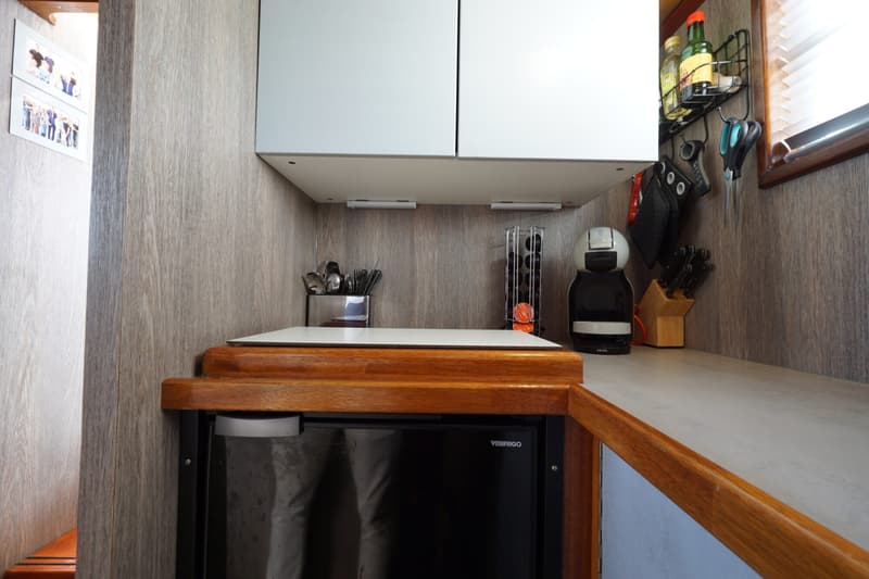 Een kajuitkeuken met een koelkast, aanrecht, bovenkasten, een koffiezetapparaat en diverse keukenbenodigdheden.
