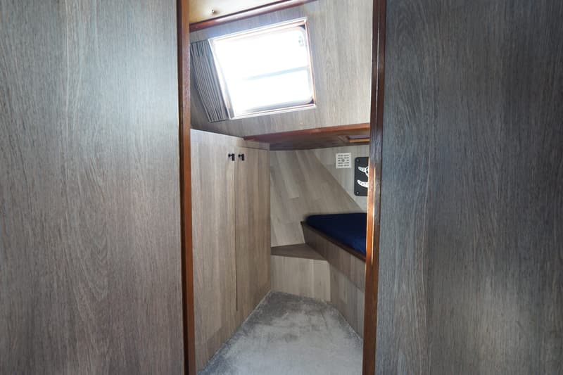 Een kajuit met een bed met blauw matras, een grijs tapijt, houten wanden en een raam met gordijn.