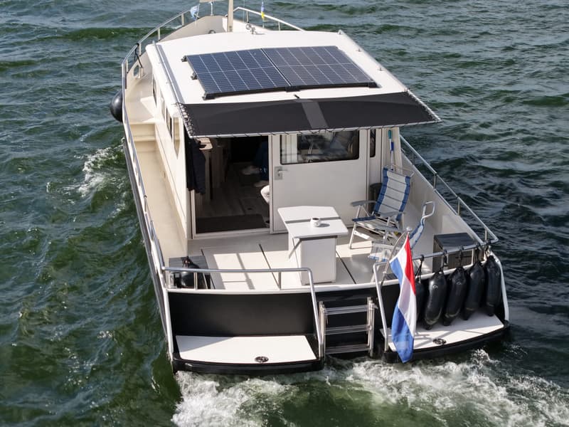 Een witte boot met zwarte accenten en zonnepanelen op het dak vaart over water, met een Nederlandse vlag aan de achterkant.