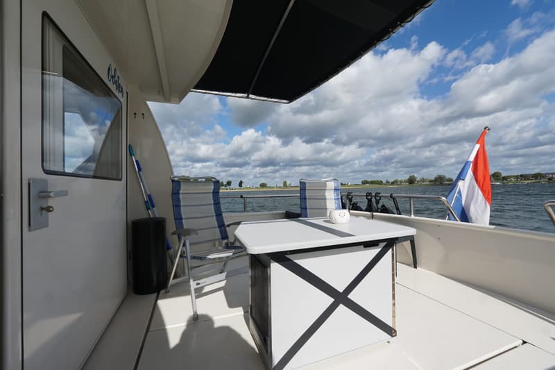 Een dek van een boot met een tafel, twee stoelen, een Nederlandse vlag en uitzicht op water en landschap.