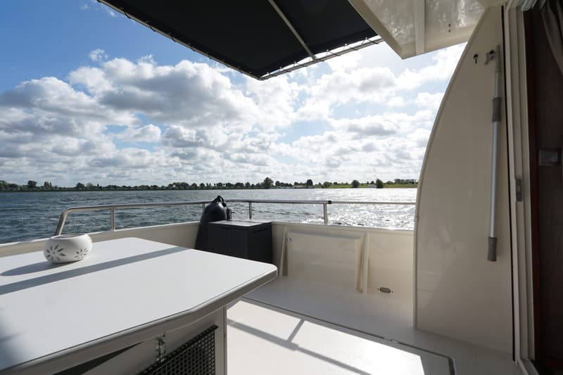 Een bootdek met een tafel en een wit ornament, met uitzicht op water en de horizon met bomen en huizen.
