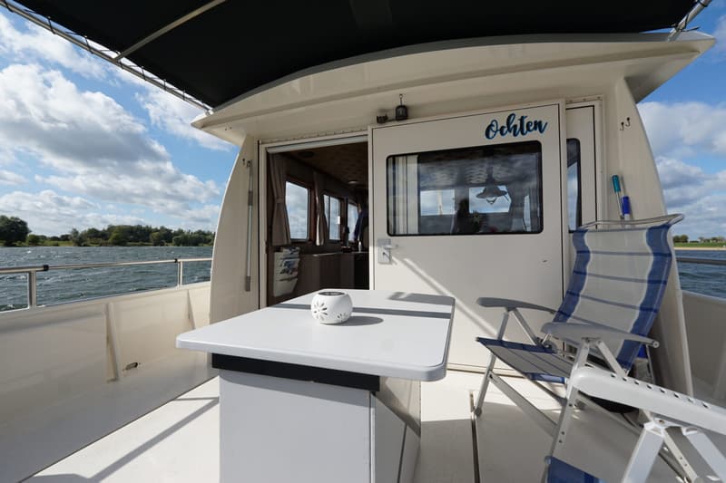 Een witte boot met een tafel en stoelen op het dek, met uitzicht op water en een beboste oever.