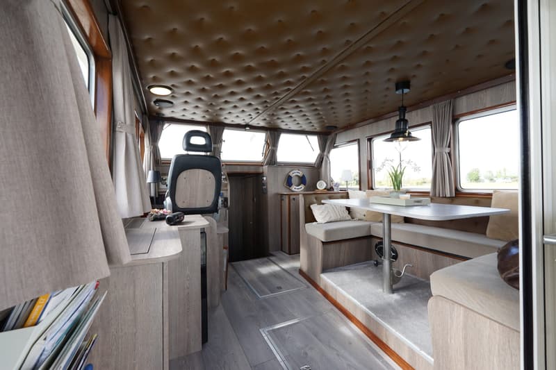 De foto toont het interieur van een boot met een stuurstoel, een tafel met zitbanken en ramen rondom.