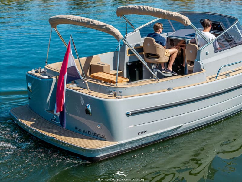 Een grijze boot met twee personen aan boord vaart op helderblauw water, met een Nederlandse vlag aan de achterkant.