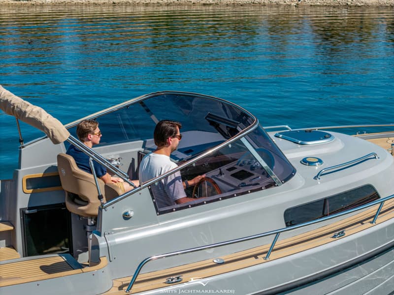 Twee mannen varen in een grijze boot met een houten stuurwiel en een beige biminitop, op blauw water.
