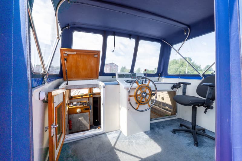 Een interieur van een boot met een stuurwiel, een bureaustoel en een open deur naar een kajuit.