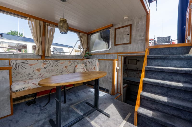 Een interieur van een boot met een zithoek, tafel, ramen met gordijnen, en een trap naar een hoger dek.