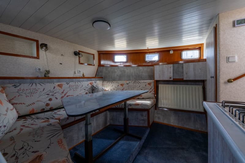 Een interieur van een boot met een zithoek, tafel, ramen, plafondlamp, en een deel van een kookplaat.