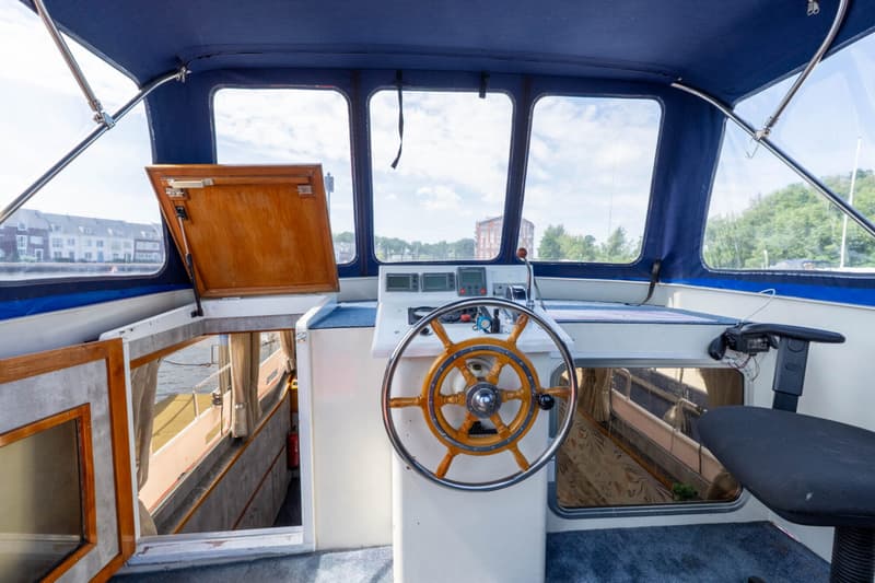 De foto toont de stuurstand van een boot met een houten stuurwiel, instrumentenpaneel en een schippersstoel.