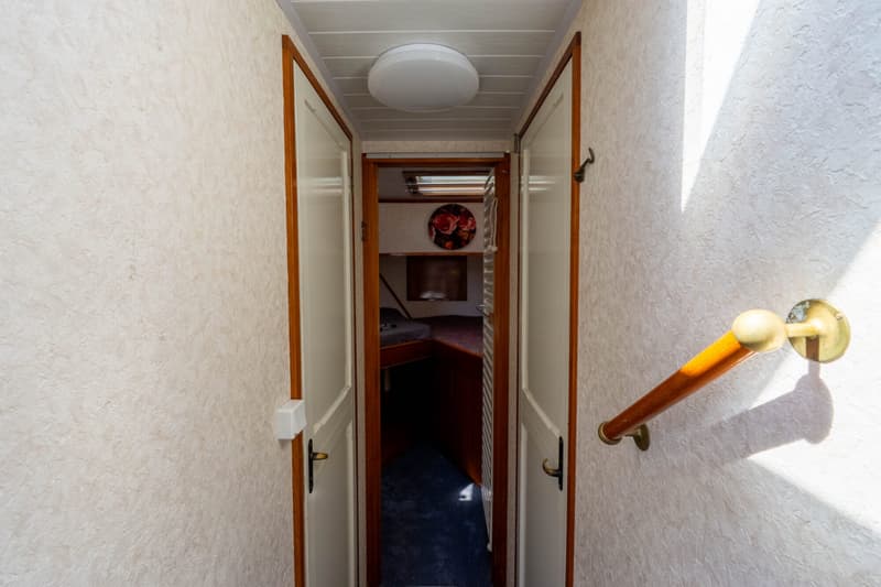 Een gang met een houten leuning aan de rechterkant, die leidt naar een open deur met een slaapruimte erachter.