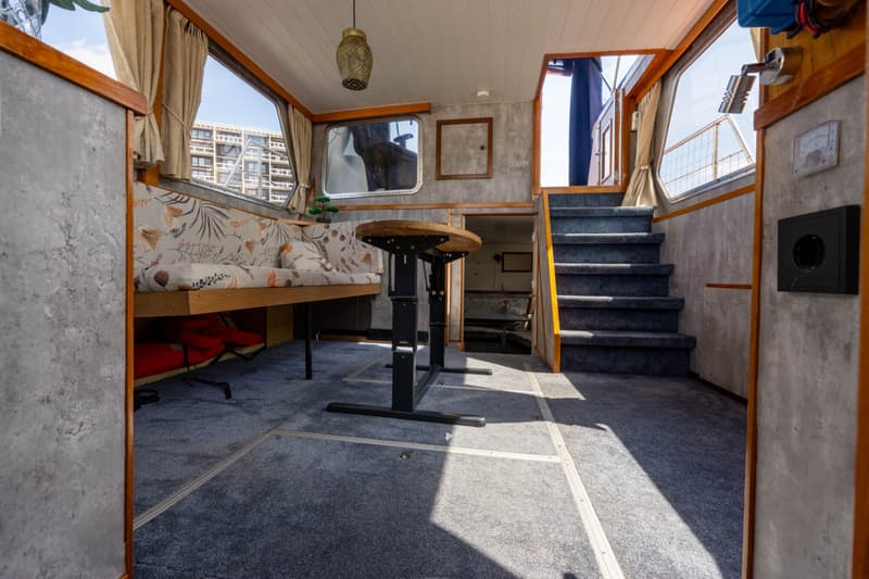 De foto toont het interieur van een boot met een zitbank, tafel, ramen, trap en tapijt.