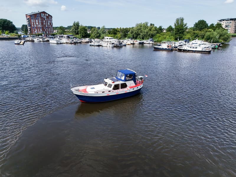 Een blauw-witte motorboot met de naam "Zuiderzee" vaart op het water, met een jachthaven en gebouwen op de achtergrond.