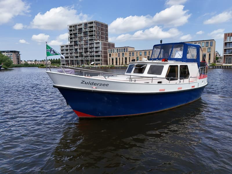 Een blauw-witte boot met de naam "Zuiderzee" ligt in het water met gebouwen op de achtergrond.