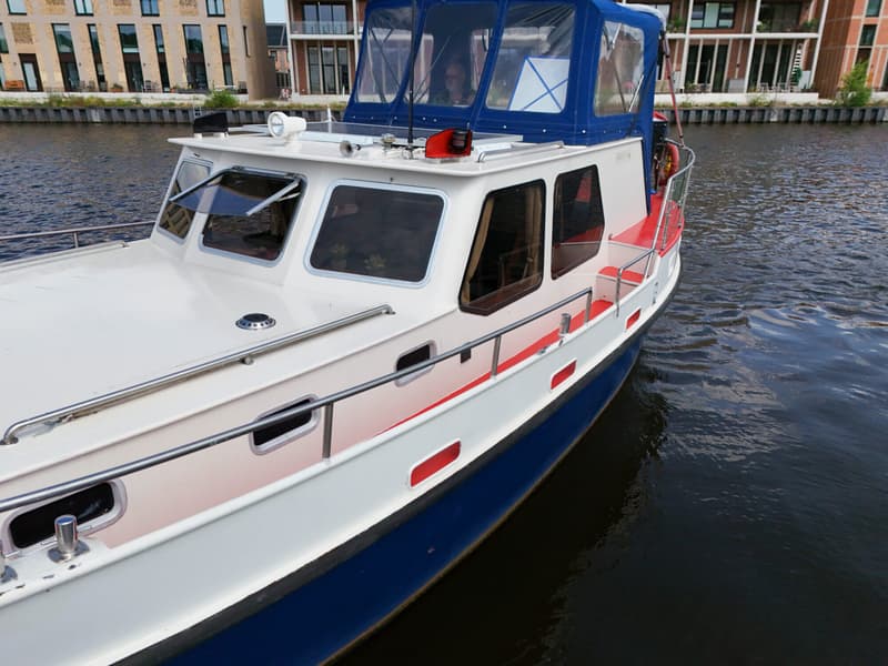 Een witte motorboot met blauwe kap en rode accenten ligt in het water, met gebouwen op de achtergrond.