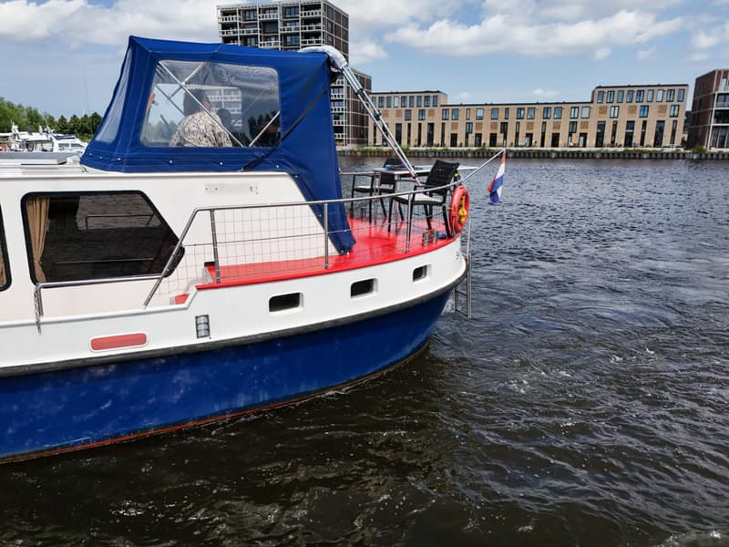 Een witte boot met blauwe romp en rode dekken vaart op het water, met gebouwen op de achtergrond.