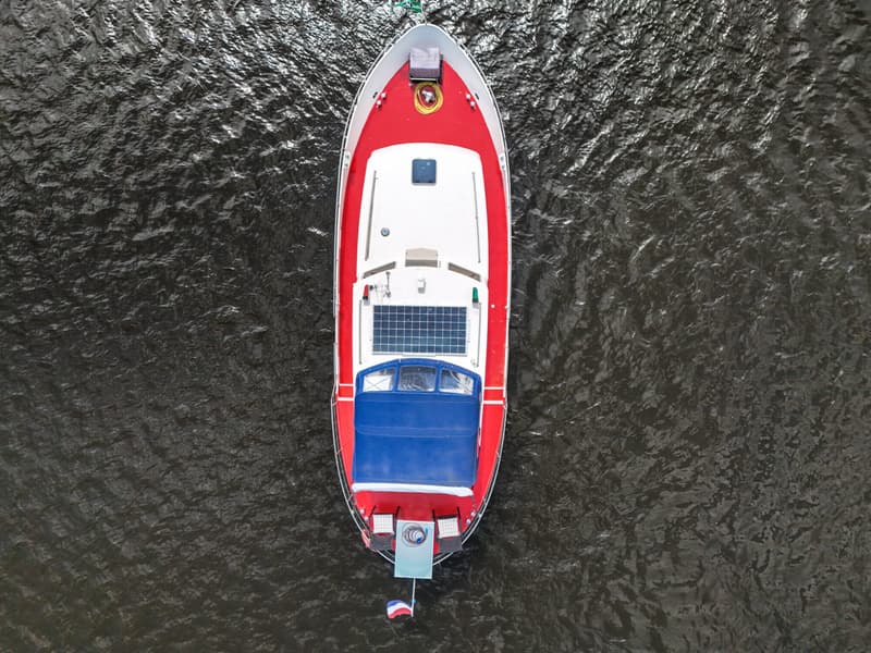 Een rood-witte boot met een blauw dekzeil en zonnepanelen, gezien van bovenaf op donker water.