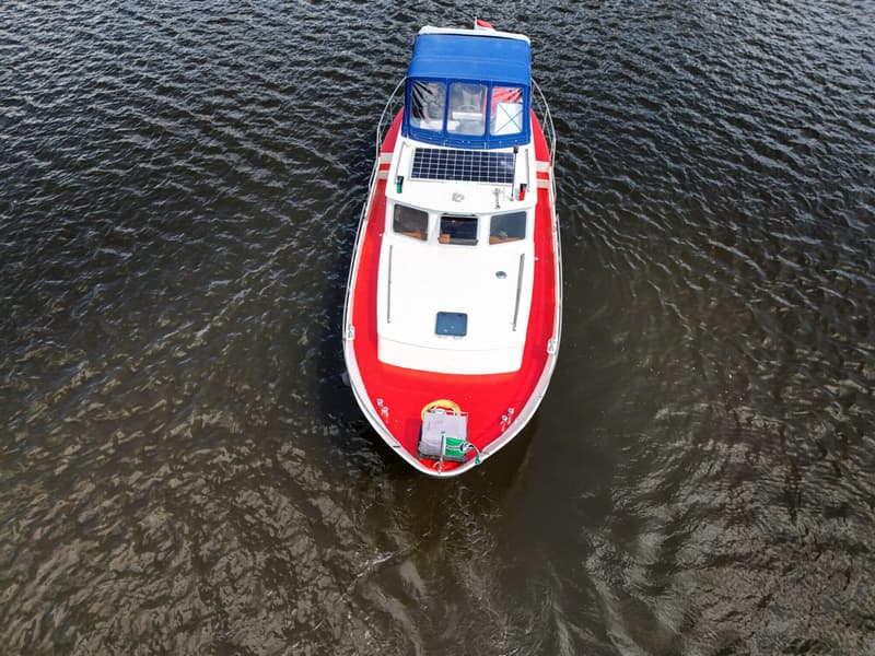 Een boot met een rode romp en een wit dek, met een blauwe kap en zonnepanelen op het dak, drijft op donker water.