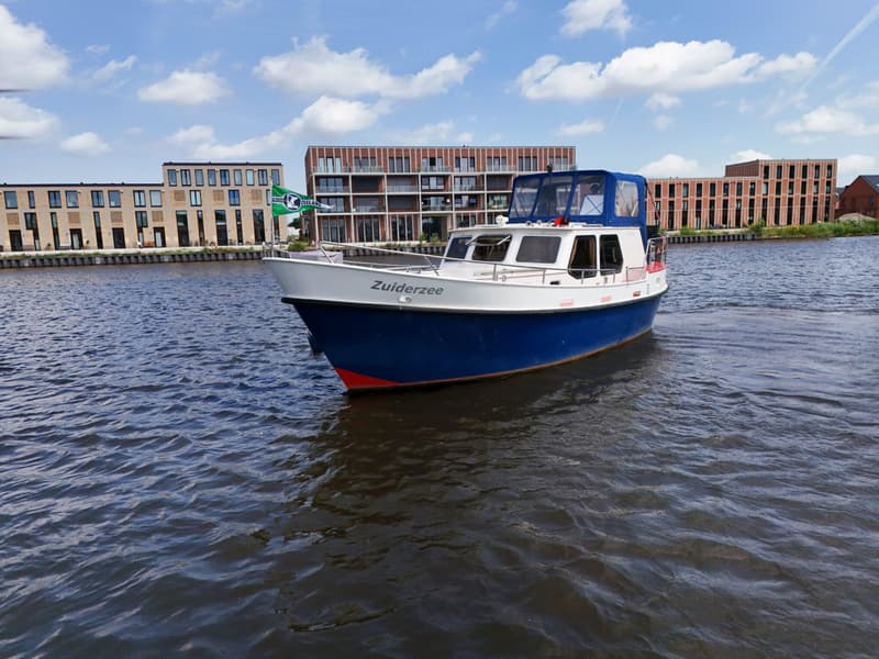 Een blauw-witte motorboot met de naam "Zuiderzee" vaart op water met gebouwen op de achtergrond.