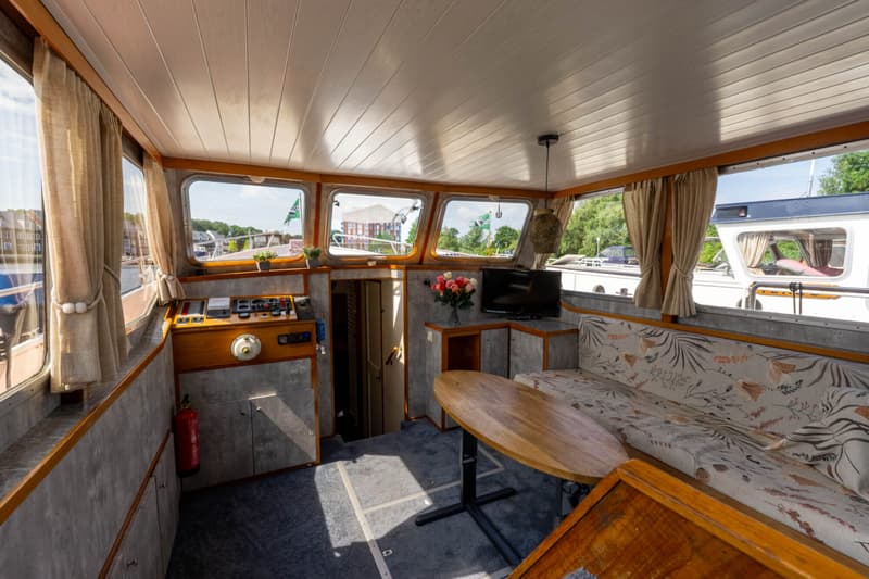 De foto toont het interieur van een boot met een stuurstand, zithoek, tafel, televisie en ramen met uitzicht.