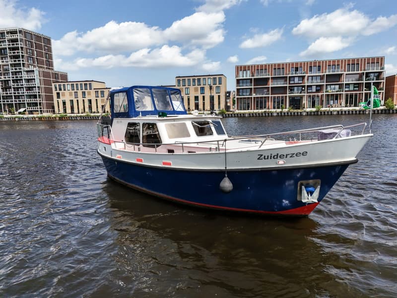 Een blauw-witte motorboot met de naam "Zuiderzee" ligt in het water voor moderne gebouwen.