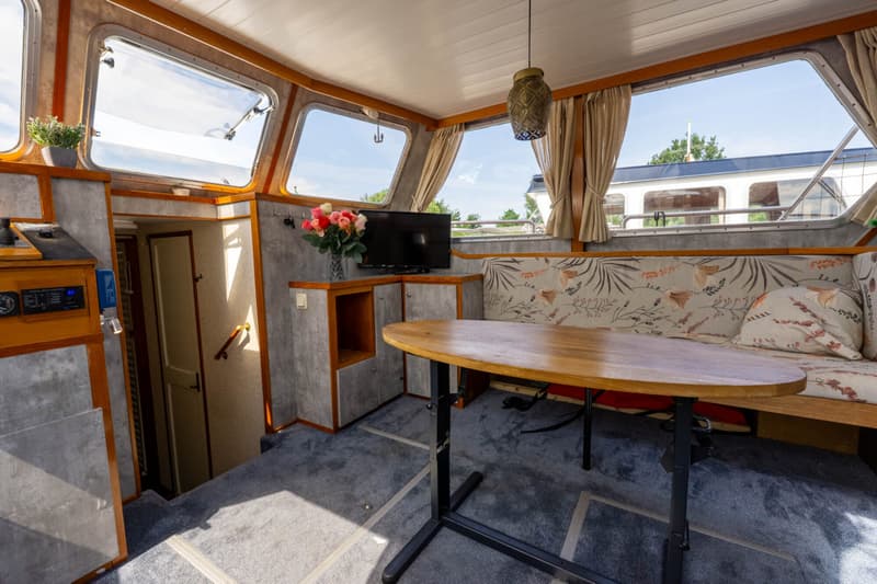 De foto toont het interieur van een boot met een houten tafel, een bank met kussens, een televisie en ramen.