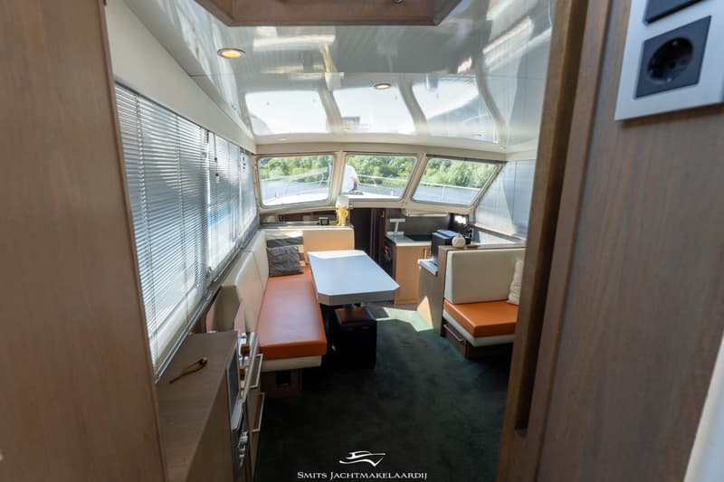 De foto toont het interieur van een boot met een zithoek, een tafel en een stuurstand, met uitzicht naar buiten.