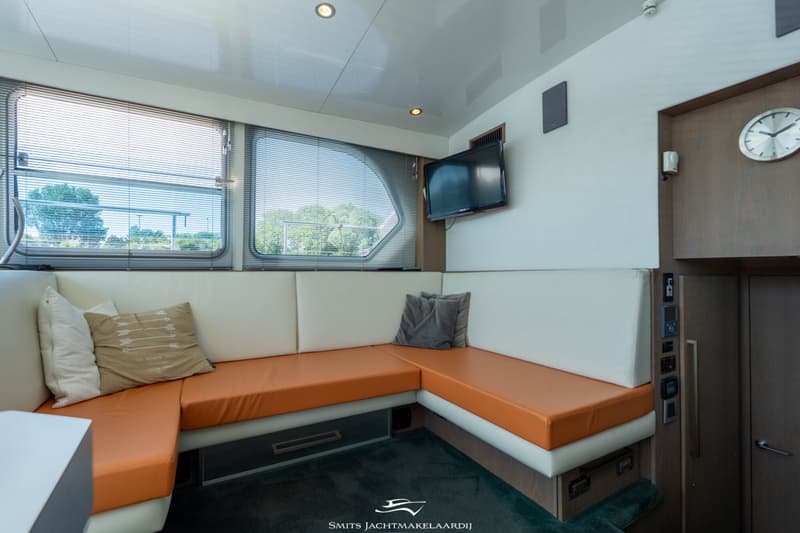 Een interieur van een boot met een L-vormige bank met oranje en witte kussens, ramen met jaloezieën, een tv en een klok.