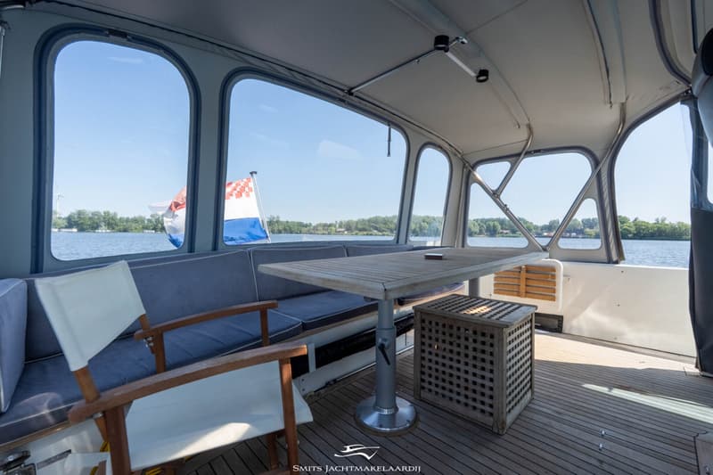 Een overdekt buitendek met een houten tafel, banken met grijze kussens en een witte stoel, met uitzicht op water en…