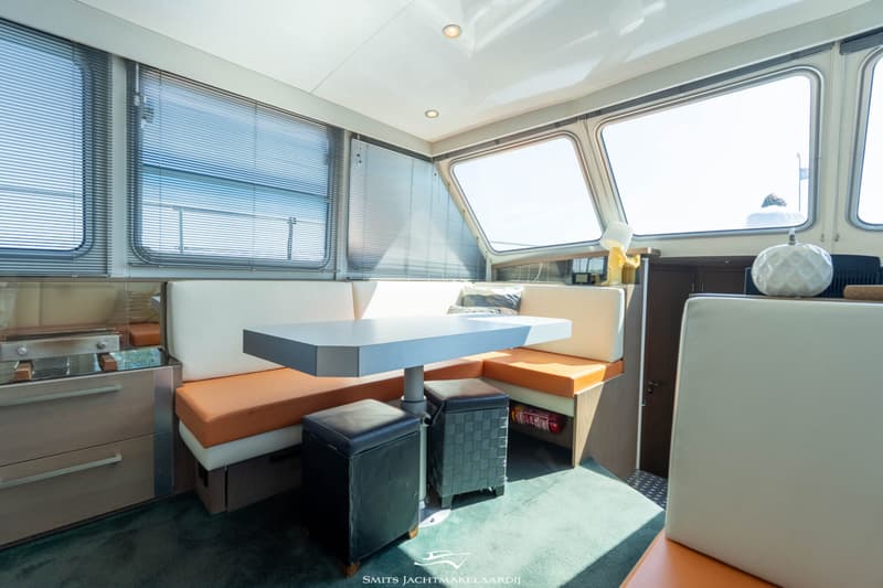 De foto toont een interieur van een boot met een eettafel, banken met oranje en witte kussens, en ramen met jaloezieën.