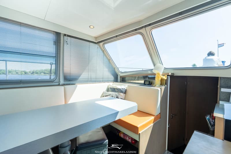 Een interieur van een boot met een witte tafel, banken met witte en oranje kussens, en grote ramen met jaloezieën.