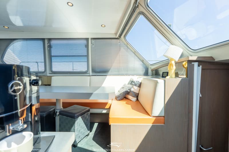 Een interieur van een boot met een zithoek, tafel, koffiezetapparaat en grote ramen met jaloezieën.