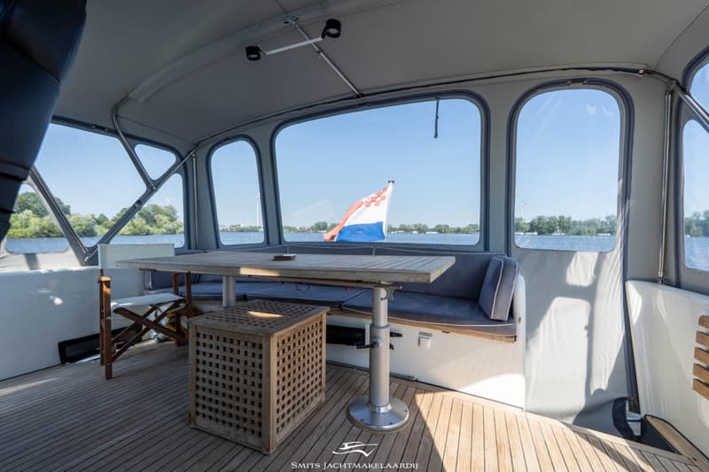 De foto toont een overdekt buitendek met een houten tafel, banken met kussens en een vlag in de verte.