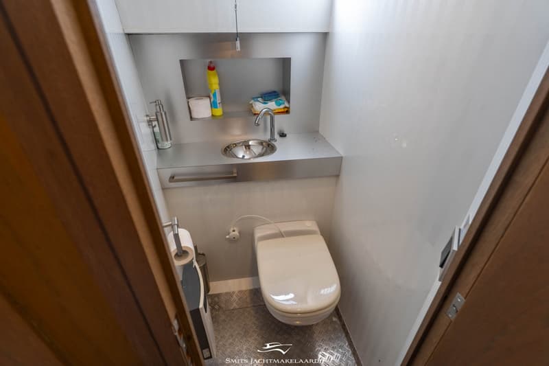 Een kleine, lichte badkamer met een toilet, wastafel en een spiegel, met schoonmaakmiddelen en toiletpapier.
