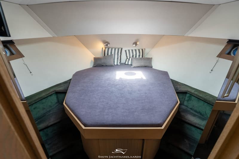 Een kajuit met een zeshoekig bed met grijs matras en kussens, twee leeslampjes en groene stoffen wandbekleding.