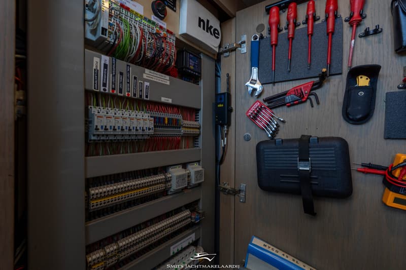 Een open paneel met elektrische bedrading, zekeringen en schakelaars, met daarnaast gereedschap aan de wand.