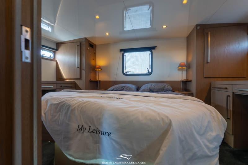 Een slaapkamer aan boord met een tweepersoonsbed, twee nachtkastjes met lampen, een raam en houten kasten.