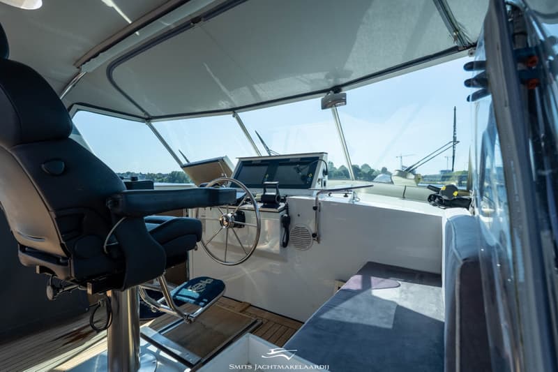 De foto toont de stuurstand van een boot met een stuurwiel, twee schermen, een kapiteinsstoel en een bankje.
