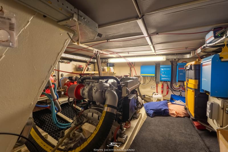 De foto toont een motorruimte met een scheepsmotor, diverse blauwe en gele elektrische componenten en bedrading.