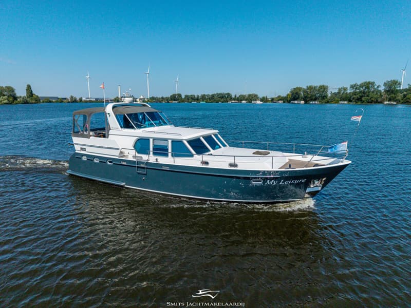 Een motorjacht met de naam "My Leisure" vaart op een waterweg met windmolens en bomen op de achtergrond.