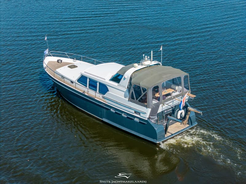 Een blauw-witte motorboot met een donkergroene bimini vaart op rustig water, gezien vanuit de lucht.