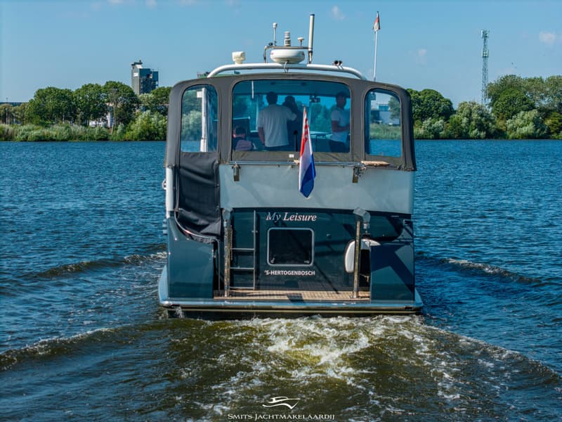 Een donkerblauwe boot met de naam "My Leisure" en " 'S-HERTOGENBOSCH" vaart over water, met drie personen aan boord.