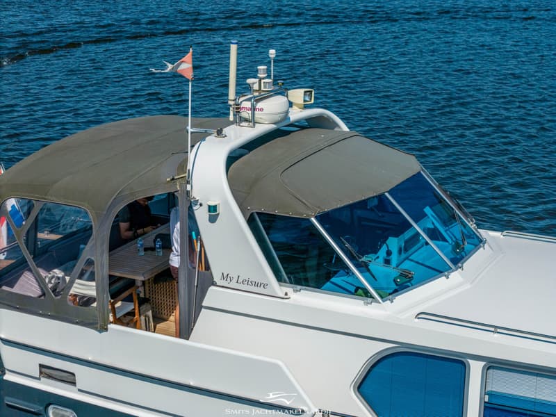 Een witte boot met een donkergroen dekzeil en de naam "My Leisure" op de zijkant, vaart op blauw water.