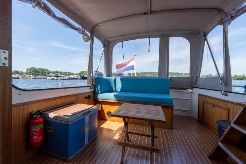 Een interieur van een boot met blauwe kussens, een houten tafel, een koelbox en een brandblusser.
